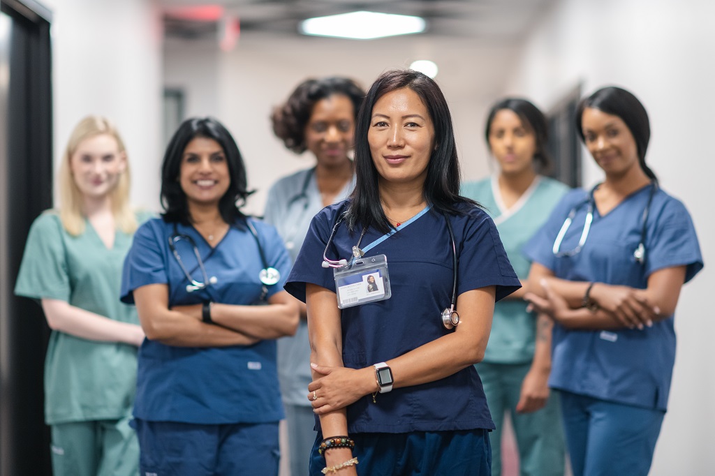 Portrait of a team of female nurses and doctors. The multi-ethnic medical professionals are standing in a corridor of a hospital. The confident group is looking directly at the camera. They are smiling with their arms crossed. A doctor of Asian descent is standing at the front of the group.